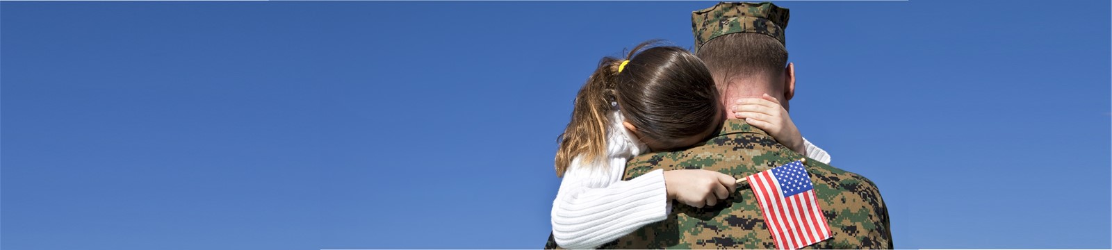 image of Marine greeting and hugging his young pony-tailed daughter who is holding an American flag after latest deployment