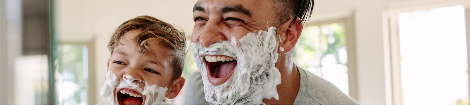 image of dark-haired dad teaching his young son how to shave. They're looking into bathroom mirror, covered with shaving cream and laughing uproariously.