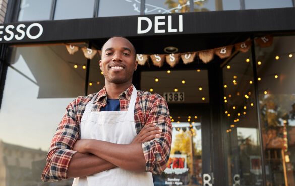 A man in a apron standing in front of a commercial building with "DELI" written on the front
