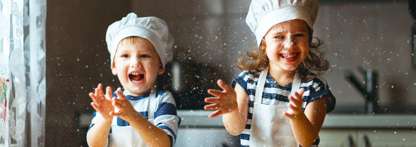 Two kids wearing aprons and chefs hats playing with baking flour